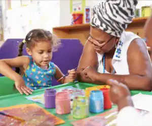 ​​Teacher sitting with child creating art with paint​