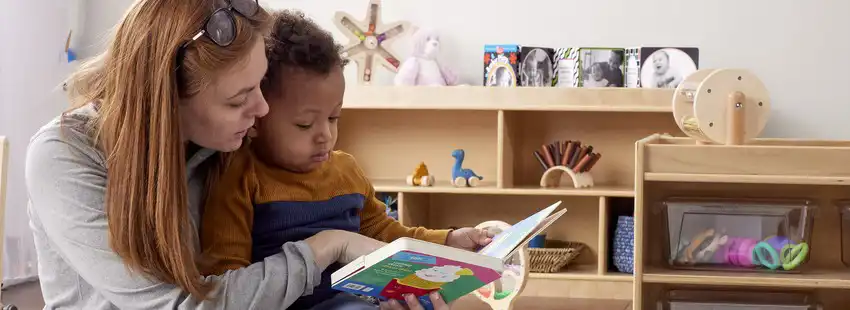 ​​Preschool teacher holding toddler while reading board book​